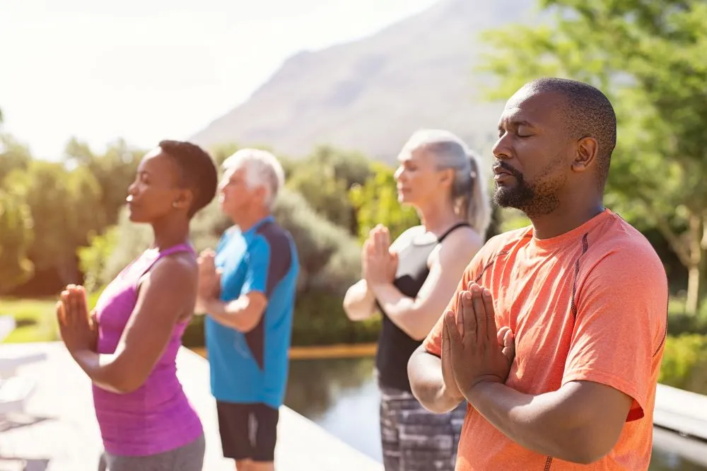 A group of men and women of different ages practices yoga and meditation outside in the sunshine